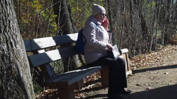 Una anciana está sentada en un banco del parque y hablando por teléfono en un caso. otoño en el parque. lleva una chaqueta, un sombrero y gafas de sol. con una bolsa en las manos — Vídeo de stock