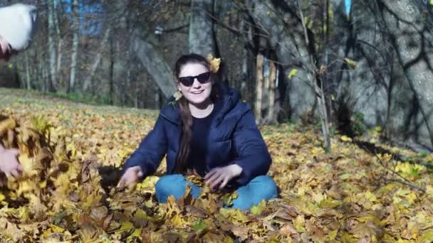Een jonge vrouw met een donkere bril zit op een tapijt van bladeren. geel blad wordt er bovenop gegooid... poserend voor een fotoshoot. — Stockvideo