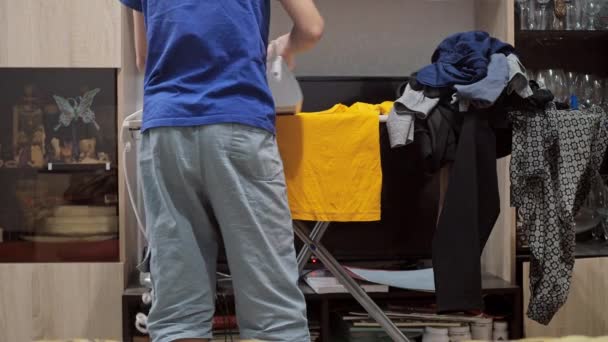 En pojke stryker en T-shirt med ett järn. hushållsarbete. Det finns en hög med skrynkliga kläder på strykbrädan. vy bakifrån — Stockvideo