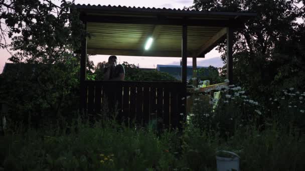 A man is standing in a gazebo. drinks a drink from a can. on a country plot. the light from the lantern. twilight. evening rest — Wideo stockowe