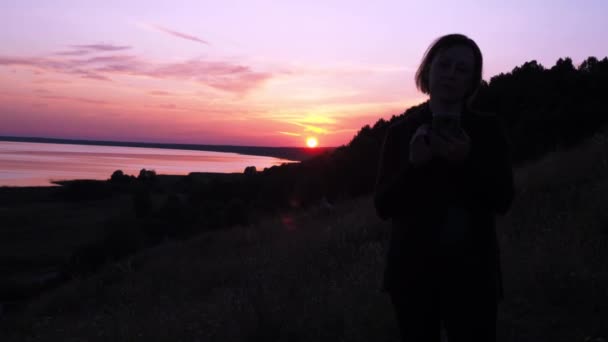 A woman stands with a phone against the background of the setting sun. red sunset in the background. hiking trips. the beauty of nature — Stockvideo