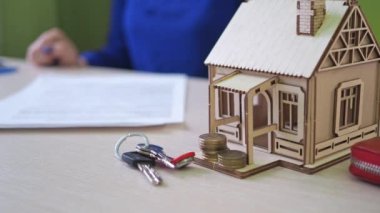 Bir kadın ev için ipotek anlaşması imzalar. Masanın üzerinde ahşap bir ev var. Arazinin anahtarları. Ön planda odaklanma. Sözleşmeyi imzalayan kadın odak dışı.