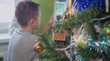 Bir genç Noel ağacının arkasında otururken tabletle oynuyor. Yeni yıl tatili. Noel ağacı odak noktasında, genç kız odak dışında.