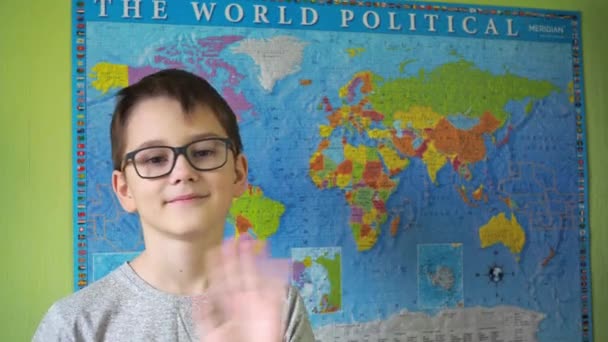 A boy with glasses waves his hand standing against the background of a political map of the world. a smile on the face of a teenager. a teenager in a T-shirt — Video Stock