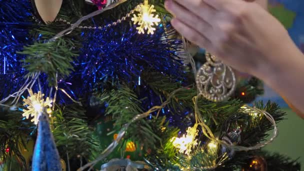 Close-up of childrens hands decorating a Christmas tree with a snowman toy against a background of bright festive lights. children decorate the Christmas tree — ストック動画