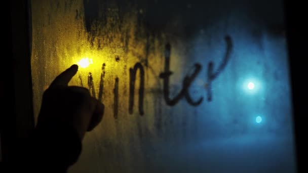 Рука пишет слово зима на туманном окне. его снег за стеклом. освещает уличный фонарь. метель ночью — стоковое видео