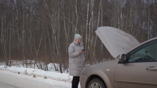 Egy nő egy lerobbant autó mellett a városon kívül, egy havas úton. veszélyes fagy. közlekedési helyzet. vontatót hívok. — Stock videók