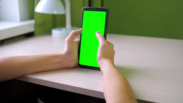 スマートフォンのクローズアップで手。指を画面にぶつけて。10代の若者が部屋のテーブルに緑の画面の電話で座っています。顔は見えません — ストック動画