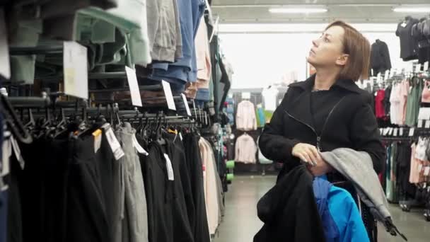 Una mujer en una tienda elige cosas. sudaderas y pantalones de chándal colgando de perchas. tienda de deportes — Vídeos de Stock