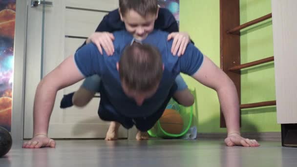 Sønnen er på sin fars ryg. armbøjninger fra gulvet, fitness derhjemme. dyrke sport sammen. karantæne i hjemmet – Stock-video