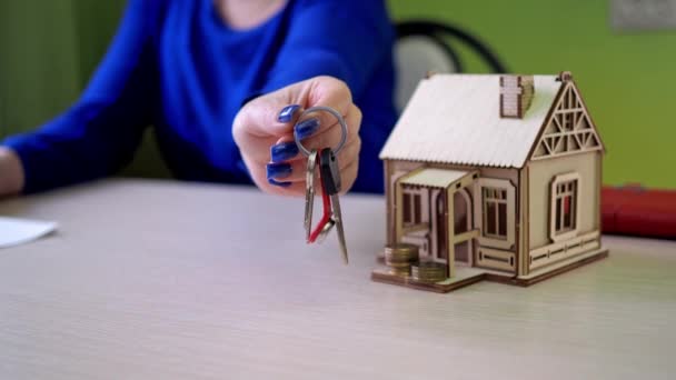 The woman holding the keys focuses on her hand in close-up. wooden house on the table. Realtor real estate agent selling real estate, make a special offer of affordable housing, moving day — Stockvideo
