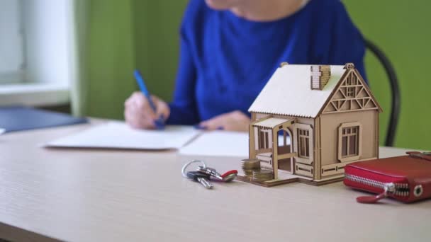 A woman signs a mortgage agreement for a house. there is a wooden house on the table, the keys to the property. foreground in focus. the woman signing the contract is out of focus — Stock Video