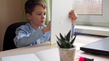 A student writes with a pencil on a tablet. a boy in a blue shirt is sitting at a table. remote activity