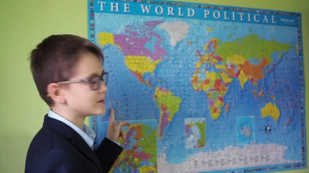 Подросток в очках и куртке указывает пальцем на страны на карте мира. мальчик в костюме и рубашке — стоковое видео