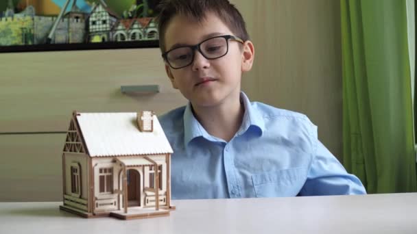 An architect boy collects a model of a house made of plywood. The boy talks about the model and shows it. After waving his hand at the camera — Vídeo de Stock