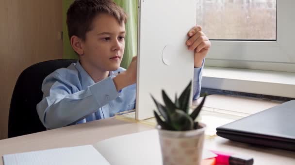 Een student schrijft met een potlood op een tablet. Een jongen in een blauw shirt zit aan een tafel. activiteit op afstand — Stockvideo