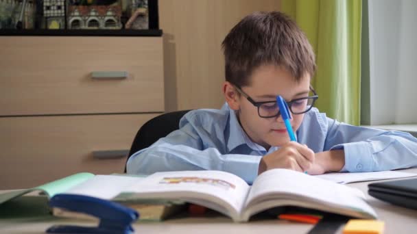 A teenager with glasses is doing homework. textbook and writing materials on the table. a desk. lessons — Wideo stockowe
