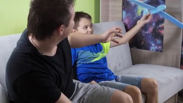 Een jongen met zijn vader op de bank spelend met een blauw vliegtuig. het vliegtuig is gemaakt van schuim. verbeelding. spelletjes voor kinderen — Stockvideo