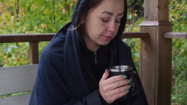Uma jovem de casaco preto está sentada a beber café quente no parque, à mesa do mirante. outono frio. Ela está constipada. corrimento nasal. aquecimento com uma bebida quente — Vídeo de Stock