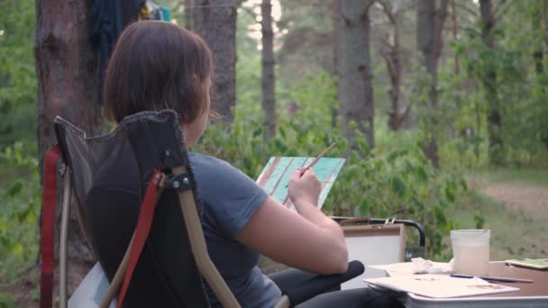 Młoda kobieta siedząca w obozowym fotelu maluje w sosnowym lesie. zdjęcie lasu — Wideo stockowe