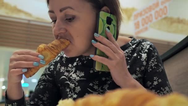 Een vrouw aan tafel eet een croissant terwijl ze telefoneert. een snelle snack bij de lunch — Stockvideo