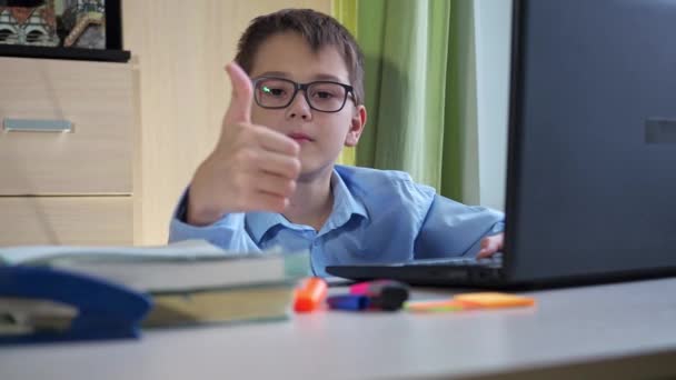 Nastolatek w niebieskiej koszuli i okularach podnosi kciuki. Siedząc przy biurku przed laptopem. nastrój pracy podczas nauki — Wideo stockowe