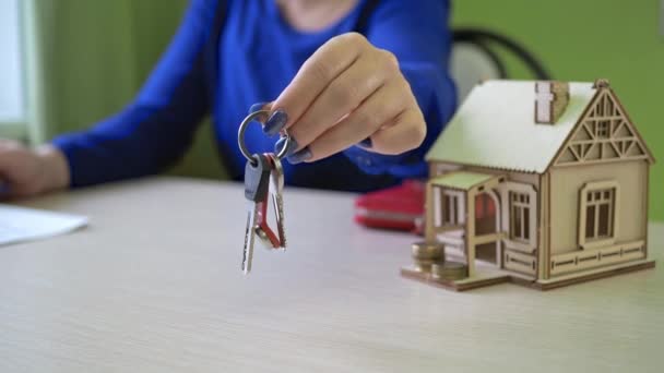 A mulher segurando as chaves se concentra em sua mão em close-up. casa de madeira na mesa. Agente imobiliário corretor que vende imóveis, fazer uma oferta especial de habitação acessível, dia em movimento, feliz — Vídeo de Stock