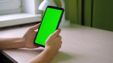 Akıllı telefonlu eller, yakın plan. Parmağıyla ekrana vuruyor. Bir genç, yeşil ekran bir telefonla odada bir masada oturuyor. Yüzü görünmüyor.