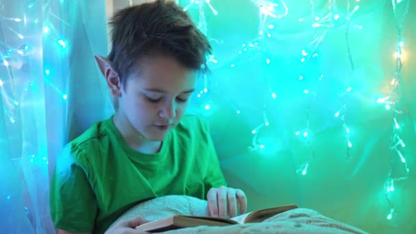 Um adolescente em um traje de duende lê um livro sentado em uma cama decorada com guirlandas. ler um livro de feitiços — Vídeo de Stock