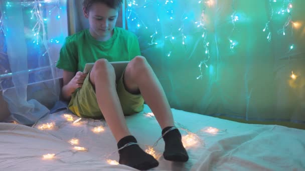The boy looks at the tablet while sitting on a bed decorated with garlands. multicolored light of lights. a teenager in a green T-shirt and shorts Stock Video
