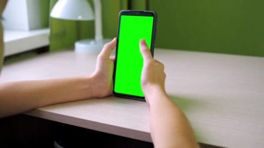 Akıllı telefonlu eller, yakın plan. Parmağını ekranda gezdiriyor. Bir genç, yeşil ekran bir telefonla odada bir masada oturuyor. Yüzü görünmüyor.