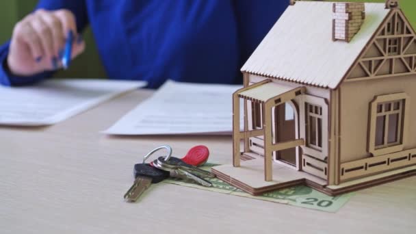 A woman signs a mortgage agreement for a house. there is a wooden house on the table, the keys to the property. foreground in focus. the woman signing the contract is out of focus — Stockvideo