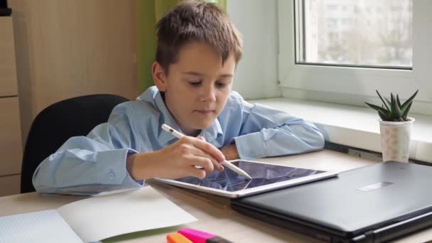 Student pisze ołówkiem na tabliczce. Chłopiec w niebieskiej koszuli siedzi przy stole. kształcenie na odległość — Wideo stockowe