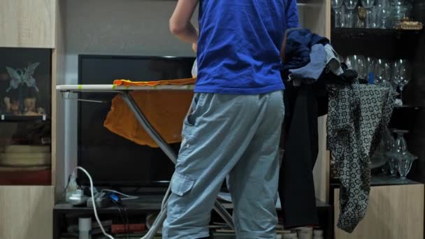 En gutt stryker en T-skjorte med et jern. husarbeid. ligger en haug med krøllede klær på strykebrettet. utsikt bakfra. uten hode – stockvideo