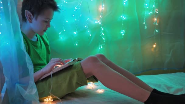 Un adolescente disfrazado de elfo lee un libro sentado en una cama a la luz de guirnaldas. decoración de Navidad de la habitación de los niños — Vídeos de Stock