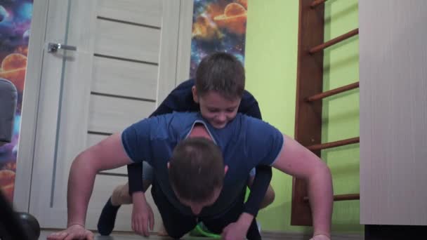 Der Sohn liegt auf dem Rücken seines Vaters. Liegestütze vom Boden, Fitness zu Hause. Sport zu Hause — Stockvideo