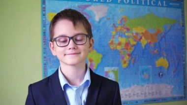 Çocuk gözlüğünü takıyor ve dünya haritasının arka planında dikilirken gözlüklerini ayarlıyor. Bir gencin yüzündeki gülümseme. Ceketli ve gömlekli bir genç.