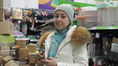 Hırdavatçıdaki bir kadın elinde telefon olan malları seçer. Beyaz şapkalı beyaz bir ceket giyiyor.