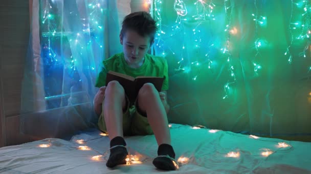Il ragazzo legge il libro ad alta voce seduto sul letto alla luce delle ghirlande. la luce multicolore delle luci. un adolescente in maglietta verde e pantaloncini — Video Stock