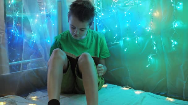 Il ragazzo legge attentamente il libro seduto sul letto alla luce delle ghirlande. la luce multicolore delle luci. un adolescente in maglietta verde e pantaloncini. lettura ad alta voce — Video Stock