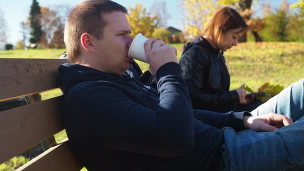 Un hombre y una mujer están sentados en un banco del parque. una mujer está hojeando una cinta en su teléfono, un hombre está bebiendo té de una taza. vacaciones familiares de otoño — Vídeo de stock