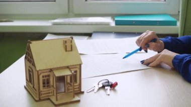 Bir kadın konut sözleşmesi imzalar. gayrimenkul satın almak. Masanın üzerinde ahşap bir ev var, evin anahtarları.