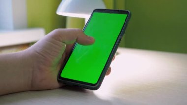 Akıllı telefonlu eller, yakın plan. Erkeklerin elleri telefondaki bandı tek eliyle çevirir. Yazı masasında bir lamba var.