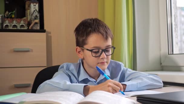 Teenager s brýlemi sedí a čte si učebnici, která sedí u stolu. psací stůl. domácí lekce. vzdálená studie. domácí prostředí — Stock video