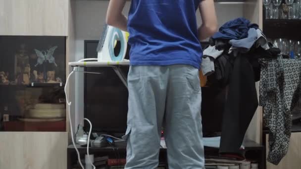 Een jongen die een T-shirt strijkt met een strijkijzer. Huishoudelijk werk. Er staat veel was op de strijkplank. zicht vanaf de achterkant — Stockvideo