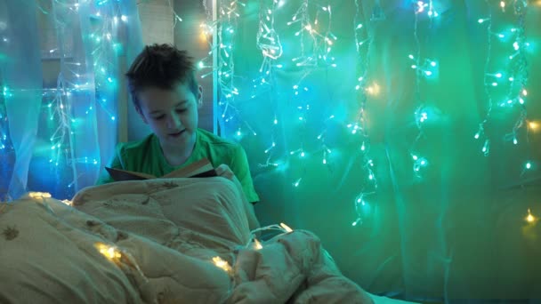 Un niño está leyendo un libro sentado en una cama decorada con guirnaldas. luz multicolor de las luces. un adolescente debajo de una manta — Vídeos de Stock