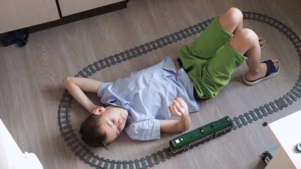 Un niño está jugando con un tren en los controles en el suelo en la habitación. Vista superior. ferrocarril de juguete. sonriendo, vuelve la cabeza hacia la locomotora en movimiento — Vídeo de stock