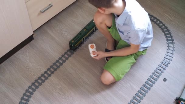Un niño con una camisa está jugando con un tren en los controles en el suelo en la habitación. Vista superior. ferrocarril de juguete — Vídeo de stock