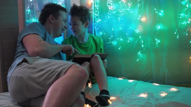 Un niño lee un libro en voz alta con su padre sentado en una cama decorada con guirnaldas de luces multicolores. crianza — Vídeos de Stock