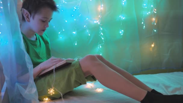 Een tiener in een elfenkostuum leest een boek op een bed in het licht van bloemenslingers. kerstversiering van de kinderkamer — Stockvideo